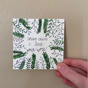 May include: A white card with green and blue foliage drawn around the edges. The card has the text "Danny Devito I love your work!" written in black ink.