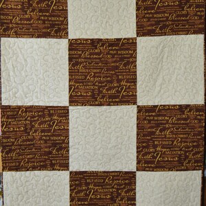 May include: A brown and white quilt with a patchwork design. Each square features a different pattern of the words "Jesus", "Faith", "Hope", "Salvation", "Rejoice", "Glory", "Wisdom", "Blessed", "Lord", "Pray", "God", "Joy", and "Christ" in a gold script font.