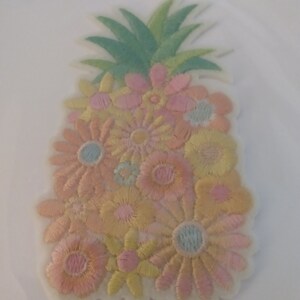 Op de afbeelding: Een witte geborduurde patch in de vorm van een ananas met een groene bovenkant en een cluster van roze, gele en perzikkleurige bloemen.