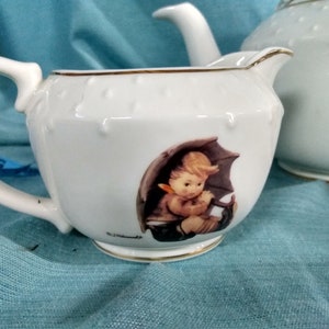 May include: White porcelain creamer with a gold rim and a printed image of a child holding an umbrella. The child is wearing a brown jacket and a red hat.