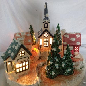 Könnte beinhalten: Eine Weihnachtsdorf-Szene aus Keramik mit einer Kirche, Häusern und Bäumen. Die Häuser haben beleuchtete Fenster und schneebedeckte Dächer. Die Bäume sind grün mit weißen Schneeakzenten.