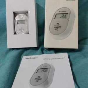 May include: A white Brookstone talking pedometer with a digital display showing 150 steps. The pedometer is packaged in a white box with the Brookstone logo and the words "talking pedometer" printed on it.