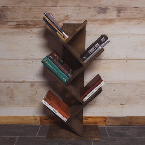 May include: A dark brown, tree-shaped bookshelf made of wood, holding several books. The bookshelf has angled shelves and is placed against a light-colored wooden wall. The books have various colored covers.