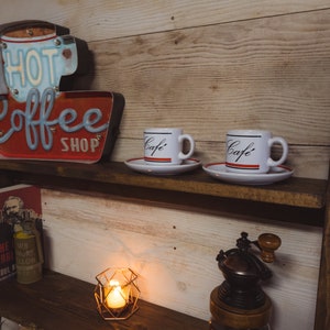 May include: A rustic wooden shelf displays coffee-themed decor. A red and blue "Hot Coffee Shop" sign sits above two white "Caf&eacute;" mugs with saucers. A book, candle, and coffee grinder complete the cozy scene.