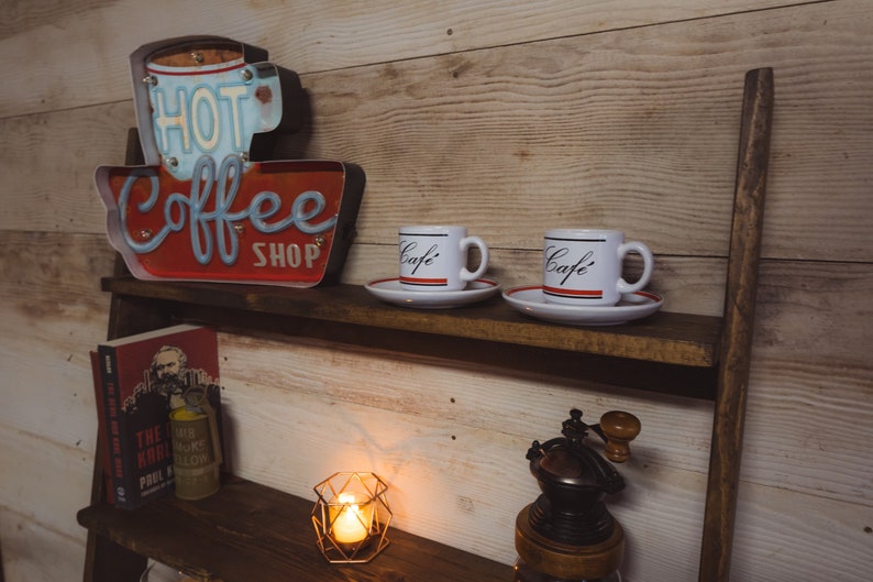 May include: A rustic wooden shelf displays coffee-themed decor. A red and blue "Hot Coffee Shop" sign sits above two white "Caf&eacute;" mugs with saucers. A book, candle, and coffee grinder complete the cozy scene.