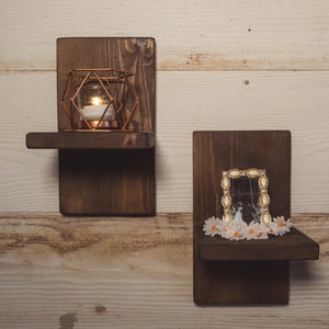 May include: Two dark stained wooden wall shelves. One shelf holds a geometric copper candle holder with a lit candle. The other shelf displays a framed photo with white flowers. The shelves are mounted on a light wood wall.