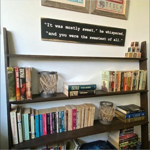 May include: A dark brown wooden bookshelf filled with books of various sizes and colors. A sign above the shelf reads "It was mostly sweet," he whispered, "and you were the sweetest of all." Decorative candle holders with rope accents are also visible.