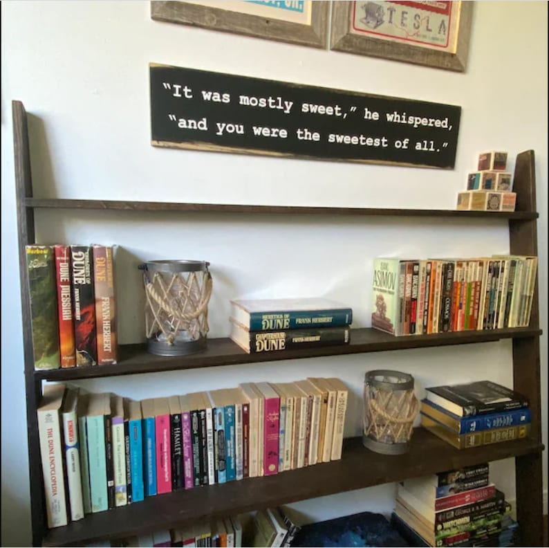 May include: A dark brown wooden bookshelf filled with books of various sizes and colors. A sign above the shelf reads "It was mostly sweet," he whispered, "and you were the sweetest of all." Decorative candle holders with rope accents are also visible.