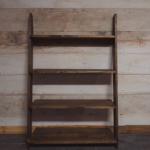 May include: A dark brown wooden leaning shelf with four shelves. The shelves are rectangular and evenly spaced. The wood has a natural grain and a rustic appearance. The background is a light-colored wooden wall.
