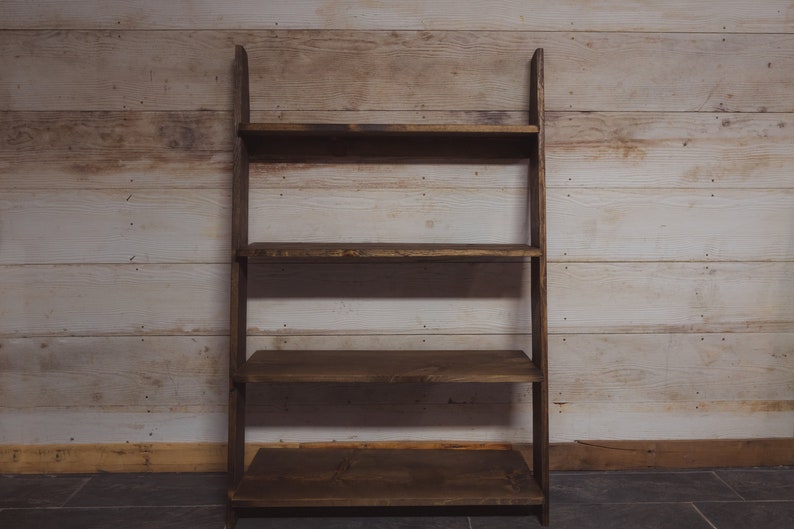 May include: A dark brown wooden leaning shelf with four shelves. The shelves are rectangular and evenly spaced. The wood has a natural grain and a rustic appearance. The background is a light-colored wooden wall.