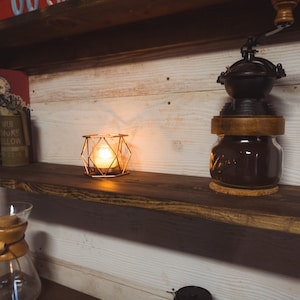 May include: A wooden shelf unit with a coffee grinder, a lit candle in a geometric cage, and books. The coffee grinder is black and sits atop a glass jar. The candle emits a warm glow.