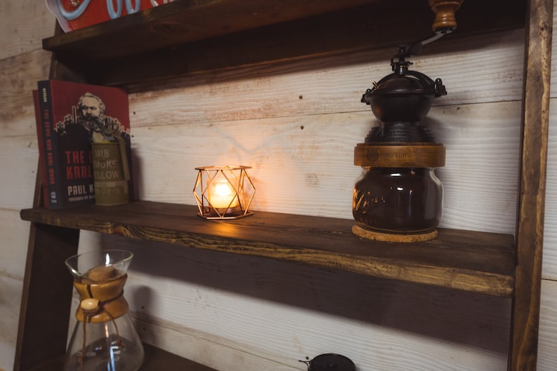 May include: A wooden shelf unit with a coffee grinder, a lit candle in a geometric cage, and books. The coffee grinder is black and sits atop a glass jar. The candle emits a warm glow.