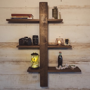 May include: A dark brown wooden wall shelf with four horizontal shelves and a vertical support. The shelves display books, a camera, a candle holder, a bottle, and other decorative items. The background is a light-coloured wooden wall.