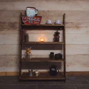 May include: A dark brown wooden shelf unit displaying coffee-themed items. The top shelf features a "Hot Coffee Shop" sign and two small coffee cups. Other shelves hold books, a candle, a coffee grinder, a camera, and a blue teapot.