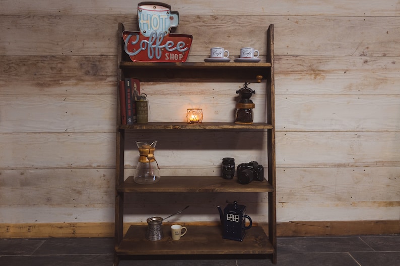 May include: A rustic, dark-stained wooden shelf unit displaying coffee-themed decor. Items include a "Hot Coffee Shop" sign, coffee cups, a coffee grinder, books, a glass coffee maker, a candle, and a blue teapot.