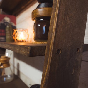 May include: A dark stained wooden shelf with a warm, inviting aesthetic. A lit candle in a glass holder, a jar, and a book are on the shelf. The shelf is part of a wooden ladder-style structure, creating a rustic display.
