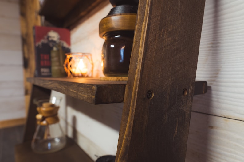 May include: A dark stained wooden shelf with a warm, inviting aesthetic. A lit candle in a glass holder, a jar, and a book are on the shelf. The shelf is part of a wooden ladder-style structure, creating a rustic display.