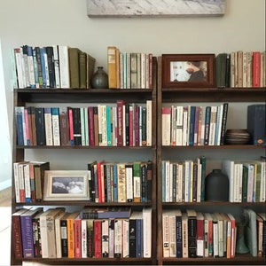 May include: A dark wooden bookshelf filled with various books of different sizes and colors. Two framed photos and two small ceramic vases are also on the shelves. The books include titles like "CHAUCER" and "Jubilee".