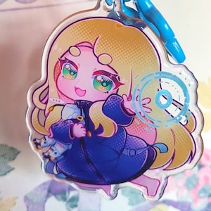May include: A clear acrylic keychain featuring a stylized illustration of a blonde character with pink skin wearing a dark blue coat. The character holds a small white object and has a glowing blue circular graphic next to them, attached to a blue ring.
