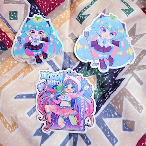 Op de afbeelding: Drie kleurrijke stickers met anime-achtige personages. Twee stickers tonen een personage met blauw haar en een microfoon. De derde sticker toont een personage met roze en blauw haar, een megafoon en Japanse tekst.