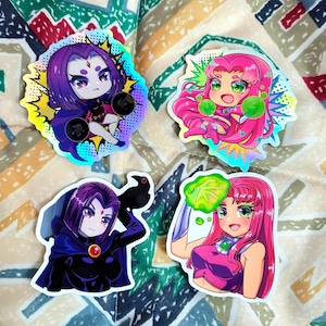 Op de afbeelding: Vier kleurrijke stickers met personages uit de animatieserie Teen Titans. De stickers tonen Raven en Starfire in verschillende poses, met levendige kleuren en holografische accenten. De stickers staan op een achtergrond van stof met patronen.