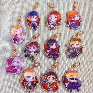 K&ouml;nnte beinhalten: Eine Sammlung von elf Acryl-Schl&uuml;sselanh&auml;ngern mit Chibi-Anime-Figuren in verschiedenen Haarfarben, Outfits und Accessoires, jeweils mit einem goldfarbenen Karabinerhaken versehen und in einem Raster auf einer strukturierten beigen Oberfl&auml;che angeordnet.