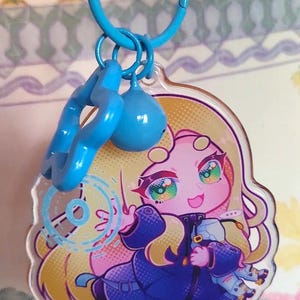 May include: A clear acrylic keychain featuring a stylized illustration of a character with long blonde hair and a dark blue coat. The character is surrounded by a light blue circular graphic. The keychain includes a blue star charm and a blue bead.