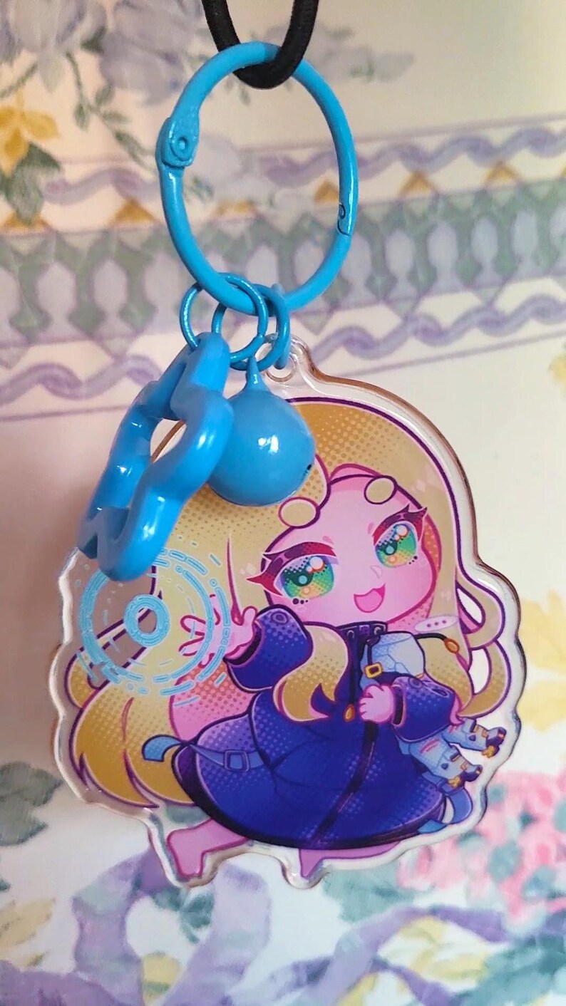 May include: A clear acrylic keychain featuring a stylized illustration of a character with long blonde hair and a dark blue coat. The character is surrounded by a light blue circular graphic. The keychain includes a blue star charm and a blue bead.