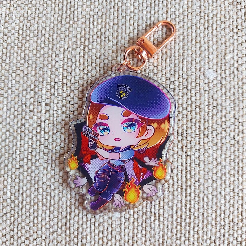 K&ouml;nnte beinhalten: Ein durchsichtiger Acryl-Schl&uuml;sselanh&auml;nger mit einer Chibi-Figur, die ein dunkelblaues Barett mit der Aufschrift S.T.A.R.S. RACCOON POLICE tr&auml;gt. Die Figur h&auml;lt eine kleine Handfeuerwaffe und ist von orangefarbenen Flammen und violetten Akzenten umgeben.