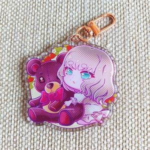 K&ouml;nnte beinhalten: Ein Schl&uuml;sselanh&auml;nger aus klarem Acryl mit einer gedruckten Illustration einer hellhaarigen Figur mit blauen Augen, die neben einem dunkelvioletten Teddyb&auml;ren mit rosa Schleife sitzt, akzentuiert durch kleine rote Erdbeergrafiken.