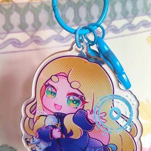 May include: A clear acrylic keychain featuring a cartoon character with long blonde hair, wearing a dark blue coat, and holding a small white object. The character has large, colorful eyes and is surrounded by a blue circular graphic element.
