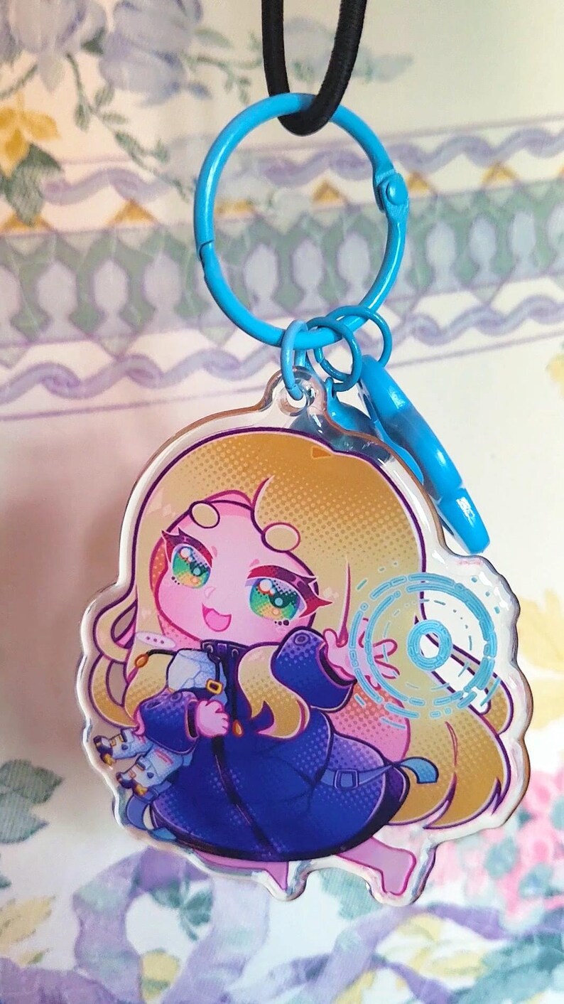 May include: A clear acrylic keychain featuring a cartoon character with long blonde hair, wearing a dark blue coat, and holding a small white object. The character has large, colorful eyes and is surrounded by a blue circular graphic element.