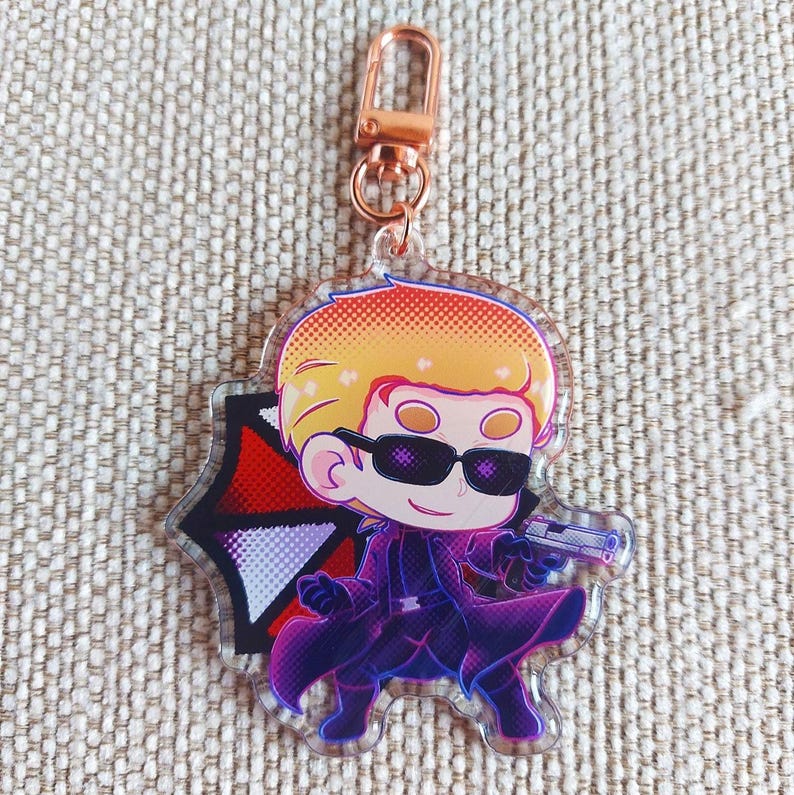 K&ouml;nnte beinhalten: Ein durchsichtiger Acryl-Schl&uuml;sselanh&auml;nger mit einer stilisierten Zeichentrickfigur mit blondem Haar, schwarzer Sonnenbrille und violettem Trenchcoat, die eine kleine silberne Pistole vor einem rot-schwarz-wei&szlig;en Regenschirm-Emblem h&auml;lt.