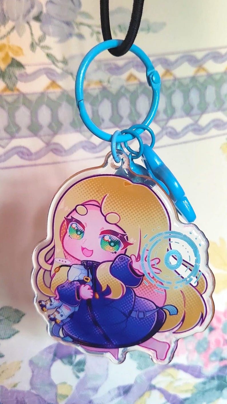 May include: A clear acrylic keychain featuring a stylized illustration of a blonde character with pink skin wearing a dark blue coat. The character holds a small white object and has a glowing blue circular graphic next to them, attached to a blue ring.