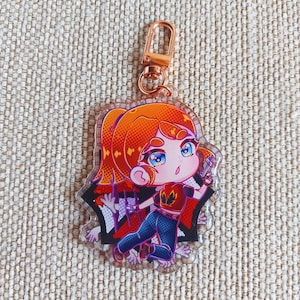 K&ouml;nnte beinhalten: Ein Schl&uuml;sselanh&auml;nger aus klarem Acryl mit einer Zeichentrickfigur mit orangefarbenen Haaren, die ein rotes T-Shirt mit Flammenemblem und eine blaue Jeans tr&auml;gt und einen kleinen grauen Gegenstand vor einem schwarzen Hintergrund h&auml;lt.