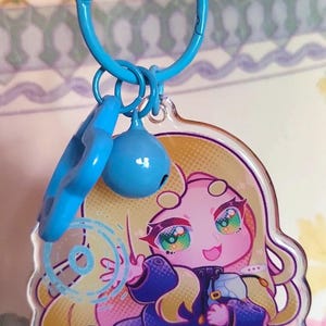 May include: A clear acrylic keychain featuring a cartoon character with long blonde hair, wearing a dark blue coat, and holding a small object. The charm is attached to a bright blue plastic ring with a matching blue bell and a star-shaped charm.