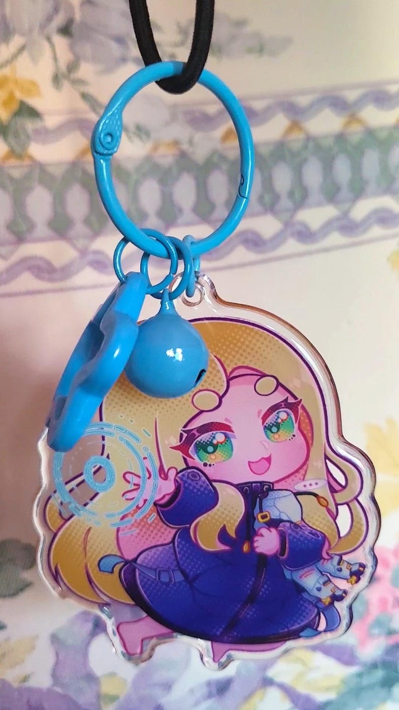 May include: A clear acrylic keychain featuring a cartoon character with long blonde hair, wearing a dark blue coat, and holding a small object. The charm is attached to a bright blue plastic ring with a matching blue bell and a star-shaped charm.