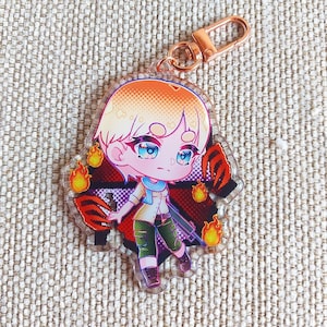 K&ouml;nnte beinhalten: Ein durchsichtiger Acryl-Schl&uuml;sselanh&auml;nger mit einer Chibi-Figur mit kurzem blondem Haar, blauen Augen, einem hellen Hemd und einer gr&uuml;nen Hose. Die Figur ist von orangefarbenen Flammen und roten Krallenspuren vor einem dunklen Hintergrund umgeben.
