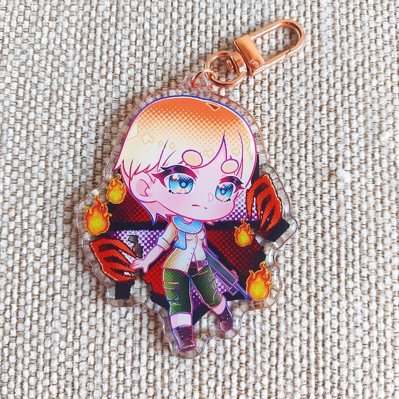 K&ouml;nnte beinhalten: Ein durchsichtiger Acryl-Schl&uuml;sselanh&auml;nger mit einer Chibi-Figur mit kurzem blondem Haar, blauen Augen, einem hellen Hemd und einer gr&uuml;nen Hose. Die Figur ist von orangefarbenen Flammen und roten Krallenspuren vor einem dunklen Hintergrund umgeben.