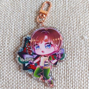 K&ouml;nnte beinhalten: Ein durchsichtiger Acryl-Schl&uuml;sselanh&auml;nger mit einer Chibi-Figur mit braunen Haaren und blauen Augen, die ein gr&uuml;n-wei&szlig;es Outfit tr&auml;gt. Die Figur h&auml;lt eine kleine graue Handfeuerwaffe vor einem dunklen, abstrakten Hintergrund mit roten und gr&uuml;nen Akzenten.