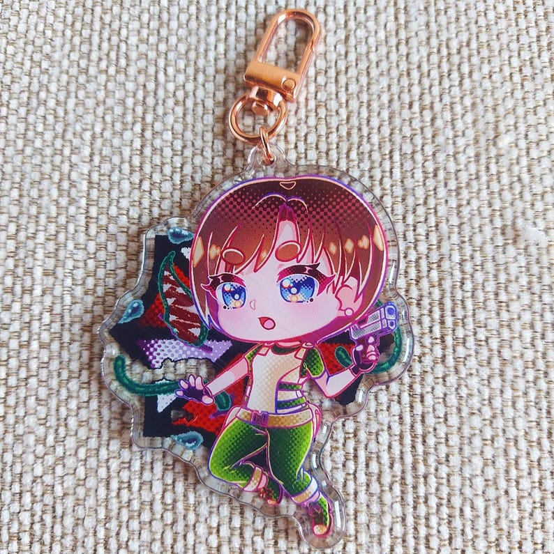 K&ouml;nnte beinhalten: Ein durchsichtiger Acryl-Schl&uuml;sselanh&auml;nger mit einer Chibi-Figur mit braunen Haaren und blauen Augen, die ein gr&uuml;n-wei&szlig;es Outfit tr&auml;gt. Die Figur h&auml;lt eine kleine graue Handfeuerwaffe vor einem dunklen, abstrakten Hintergrund mit roten und gr&uuml;nen Akzenten.