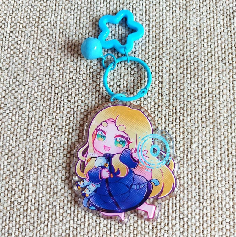 May include: A clear acrylic keychain featuring a cartoon character with long blonde hair, wearing a dark blue jacket, and holding a small object. The keychain includes a light blue star-shaped clasp, a small round bell, and a circular ring.
