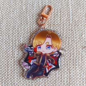 K&ouml;nnte beinhalten: Ein Acryl-Schl&uuml;sselanh&auml;nger mit einer Chibi-Figur mit blondem Haar, die eine braune Jacke und eine dunkle Hose tr&auml;gt und eine graue Handfeuerwaffe h&auml;lt. Die Figur ist vor einem schwarz-roten Hintergrund mit einem ros&eacute;goldenen Karabinerhaken.