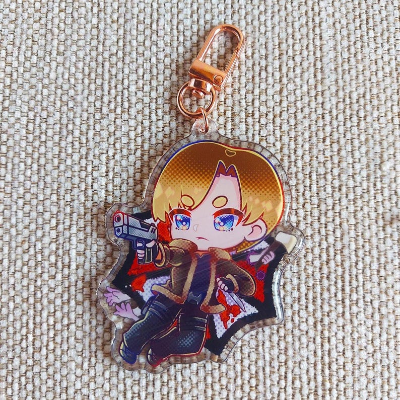 K&ouml;nnte beinhalten: Ein Acryl-Schl&uuml;sselanh&auml;nger mit einer Chibi-Figur mit blondem Haar, die eine braune Jacke und eine dunkle Hose tr&auml;gt und eine graue Handfeuerwaffe h&auml;lt. Die Figur ist vor einem schwarz-roten Hintergrund mit einem ros&eacute;goldenen Karabinerhaken.