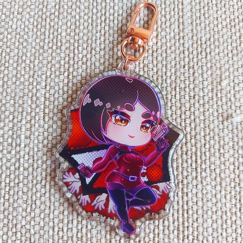 K&ouml;nnte beinhalten: Ein Schl&uuml;sselanh&auml;nger aus klarem Acryl mit einer Chibi-Figur mit kurzem dunklem Haar, die einen dunkelroten Anzug tr&auml;gt und eine kleine Handfeuerwaffe h&auml;lt. Das Hintergrunddesign zeigt rote und schwarze geometrische Muster mit wei&szlig;en Handumrissen.