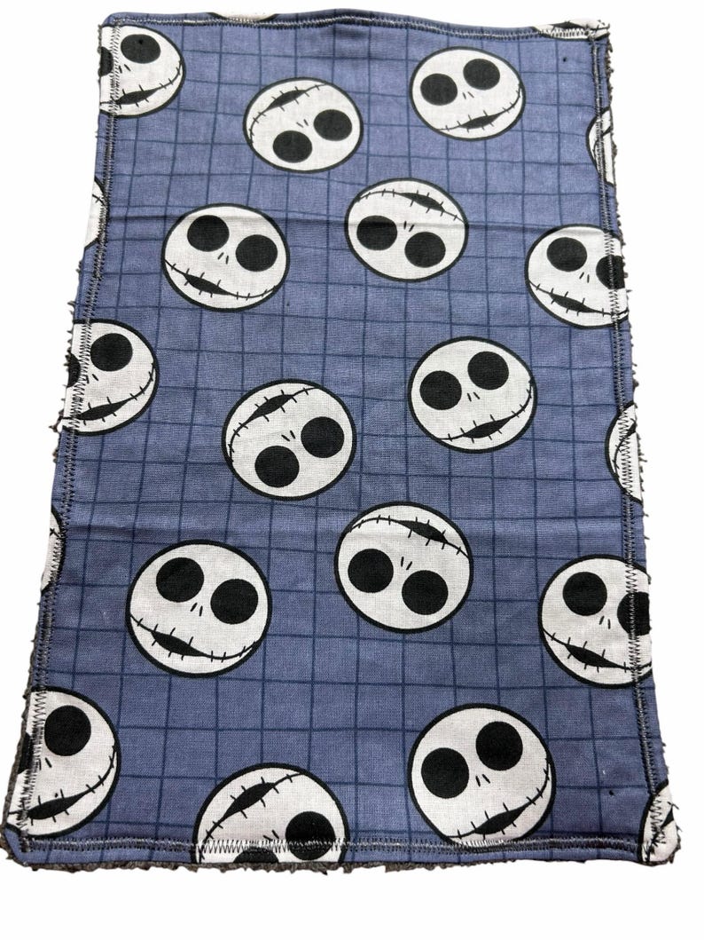 May include: A blue cloth featuring a grid pattern and repeated white Jack Skellington faces. The edges of the cloth are frayed. The Jack Skellington faces have black eyes and stitched smiles.