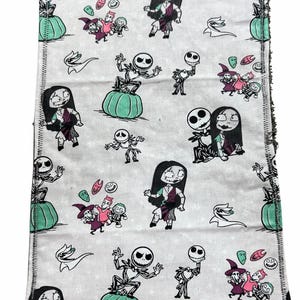 May include: A rectangular, light grey fabric featuring a repeating pattern of characters from The Nightmare Before Christmas. The design includes Jack Skellington, Sally, and other characters, along with pumpkins and other Halloween-themed elements. The edges have a black stitched border.