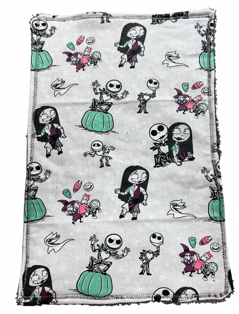 May include: A rectangular, light grey fabric featuring a repeating pattern of characters from The Nightmare Before Christmas. The design includes Jack Skellington, Sally, and other characters, along with pumpkins and other Halloween-themed elements. The edges have a black stitched border.