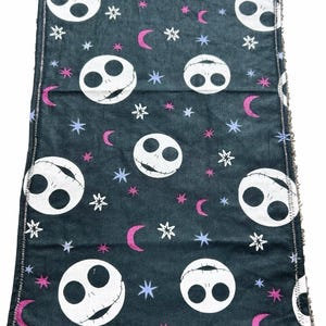 May include: A dark grey cloth with a repeating pattern of white Jack Skellington faces, pink crescent moons, white stars, and blue stars. The edges are finished with a contrasting stitch.