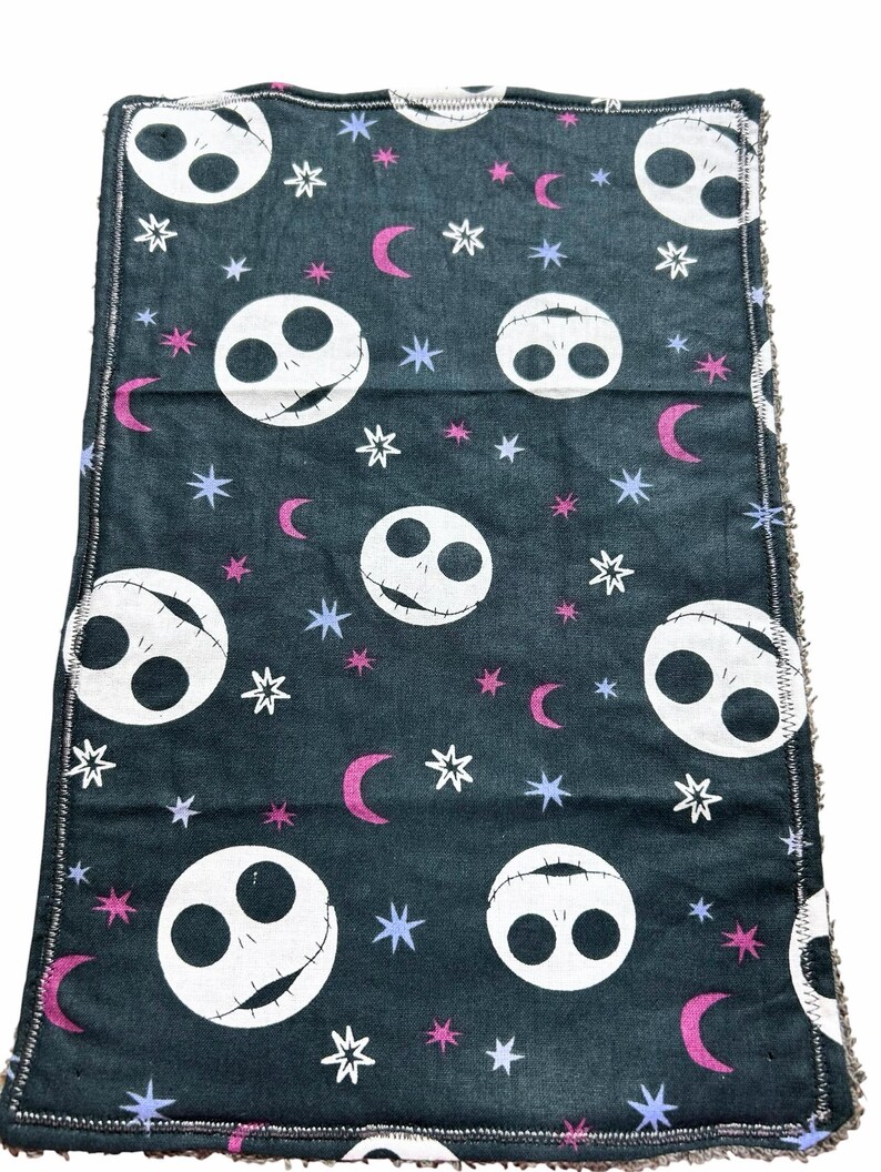May include: A dark grey cloth with a repeating pattern of white Jack Skellington faces, pink crescent moons, white stars, and blue stars. The edges are finished with a contrasting stitch.